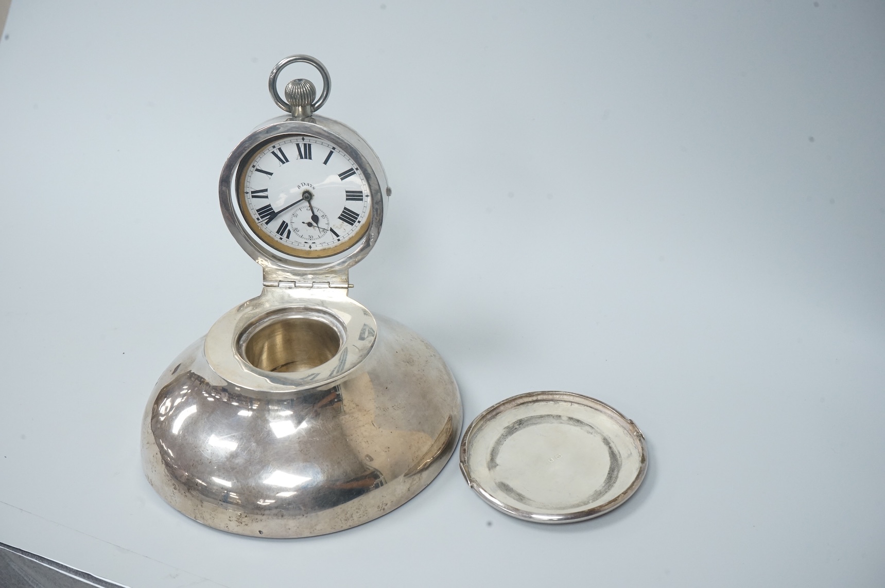 An Edwardian silver circular inkwell, the hinged cover inset with an 8-day open faced pocket watch, with Roman dial and subsidiary seconds, makers mark rubbed, Saunders & Shepherd?, Birmingham, 1907, diameter of the base
